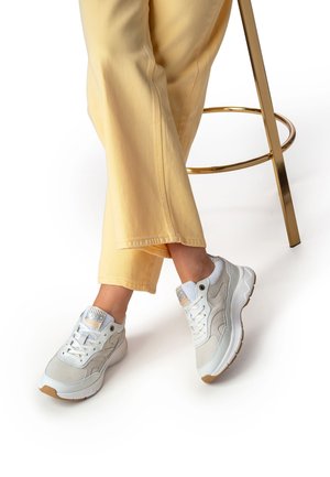 Witte sneaker met mesh- en leren panelen, lichtgrijze accenten, afgeronde neus, veterdetails en een gestructureerde zool. Gecombineerd met lichtgele broek.