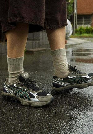 Scarpe da corsa nere e argento con dettagli verdi, lacci e suole texturizzate, abbinate a calzini a coste grigi chiari, poggiate su un pavimento bagnato.