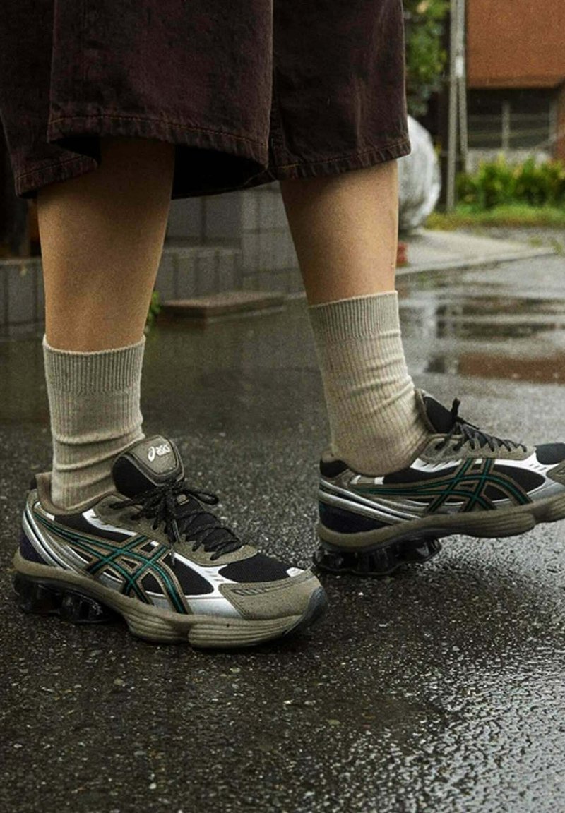 Scarpe da corsa nere e argento con dettagli verdi, lacci e suole texturizzate, abbinate a calzini a coste grigi chiari, poggiate su un pavimento bagnato.