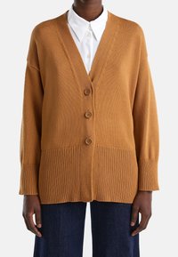 Brun stickad kofta med V-ringning, med ribbade detaljer, tre knappar och sänkt axel. Bärs över en vit skjorta med krage.