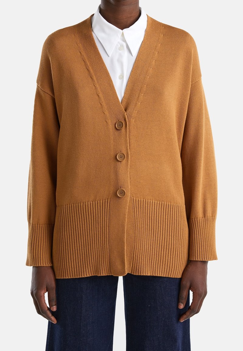 Brun stickad kofta med V-ringning, med ribbade detaljer, tre knappar och sänkt axel. Bärs över en vit skjorta med krage.