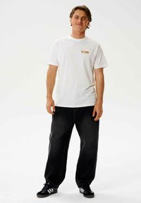 Hvid kortærmet t-shirt med et lille orange logo, parret med løstsiddende sorte jeans og sorte sneakers med hvide striber.