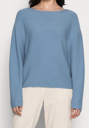 Femme portant un pull en tricot bleu clair à manches longues et un pantalon beige, vue du cou aux hanches sur un fond uni.