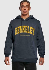 Marineblå hettegenser med gult "BERKELEY"-trykk og "University of California" under. Har en lomme foran og justerbar hette med snøring.
