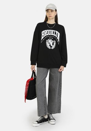 Felpa nera con stampa "BROOKLYN", jeans larghi grigi, sneakers nere e cappellino grigio chiaro. Borsa tote nera con un accento in bandana rossa.