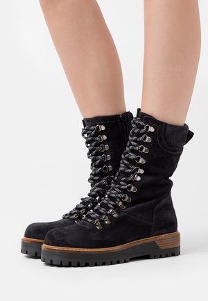 Botas de tobillo de ante negro con ojales de metal, cordones gruesos negros, un zipper lateral y una suela de goma gruesa con un acento de madera.