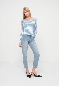 Μπλούζα με μπλε και λευκές ρίγες, χωρίς ώμους, ανοιχτόχρωμο μπλε τζιν, μαύρα σανδάλια slingback με λεπτομέρεια φιόγκου. Το μοντέλο στέκεται σε ουδέτερο φόντο.