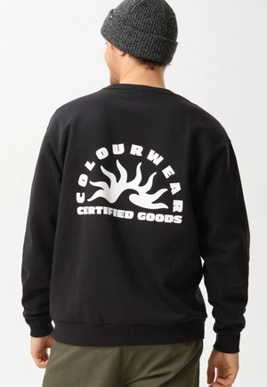 Man draagt een zwarte sweatshirt met een wit zonlogo en de tekst "Colourwear Certified Goods" op de achterkant, en een donkergrijze gebreide muts.