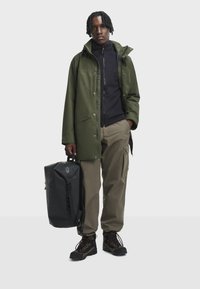 Zelená nepromokavá bunda přes černou fleecovou mikinu na zip, béžové cargo kalhoty, černé turistické boty a černý batoh s bočními popruhy, vystavené na bílém pozadí.