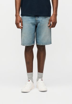 Bărbat purtând pantaloni scurți din denim albastru deschis, șosete gri, adidași albi și o cămașă de culoare albastru închis, stând în picioare pe un fundal alb.