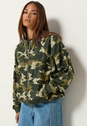 Femme aux longs cheveux bruns portant une veste zippée camouflage verte et beige et un jean bleu, appuyée contre un mur blanc.