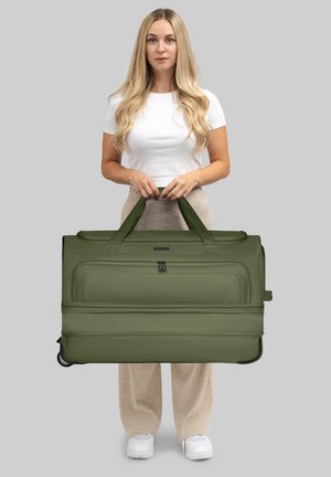 Donna con lunghi capelli biondi, che indossa una maglietta bianca e pantaloni beige, mentre tiene un grande borsone verde oliva con ruote e una tasca frontale.
