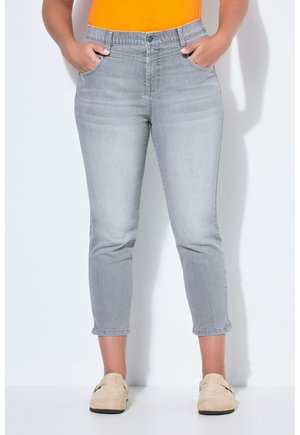 Frau trägt hellgraue, verkürzte Jeans mit Vordertaschen, kombiniert mit beigen Slippern und einem orangen Oberteil, steht vor einer weißen Wand.