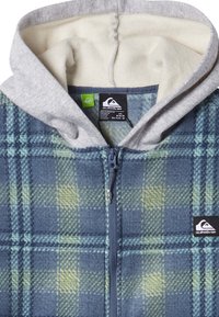 Fleece zip-up huppari, jossa on harmaa huppu. Sinivihreä ruudullinen kuvio, pehmeä rakenne, etuvetoketju ja brändilogo-tarra alhaalla vasemmalla.