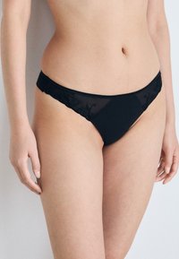 Culotte noire en dentelle avec des panneaux latéraux en tulle transparent, broderie florale à la taille, et corps en tissu opaque et lisse.