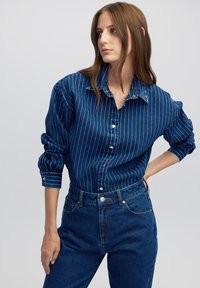 Dunkelblaue Jeanshemd mit weißen vertikalen Streifen, Knopfleiste vorne und hochgerollten Ärmeln. Kombiniert mit hoch taillierten blauen Jeans.