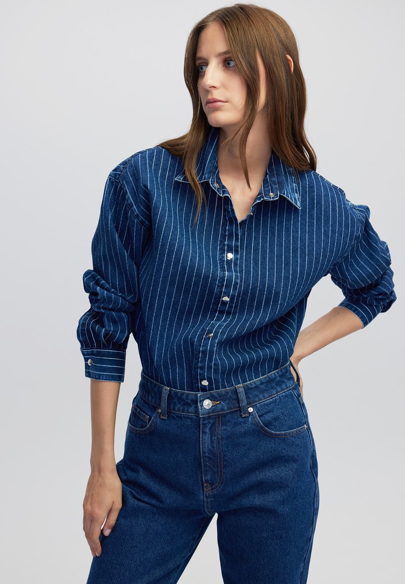 Camisa de denim azul oscuro con rayas verticales blancas, con botones en la parte delantera y mangas enrolladas. Combinada con unos pantalones de denim azul de cintura alta.