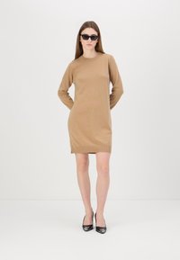 Abito lungo in maglia color beige con maniche lunghe, scollatura a girocollo, spacchi laterali e silhouette aderente, abbinato a décolleté nere a punta e occhiali da sole.