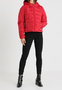 Giacca a vento rossa e gonfia con cappuccio, dotata di una zip frontale e cuciture orizzontali. Abbinata a pantaloni neri slim-fit e stivaletti lucidi alla caviglia.