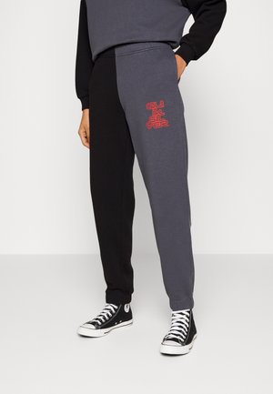 Persona con pantalones deportivos divididos negros y gris oscuro con logo rojo de Quiksilver en el muslo y zapatillas negras de caña alta con cordones blancos.