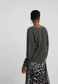 Übergroßer dunkelgrüner Strickpullover mit Rippstruktur, kombiniert mit einem fließenden Rock in Schwarz-Weiß mit Tiermuster, von hinten sichtbar.