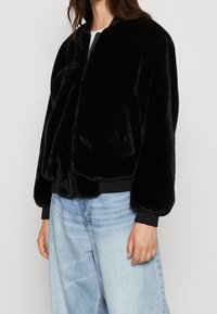 Sort faux fur bomberjakke med lynlås foran, ribbede manchetter og kant. Båret over en hvid top og parret med lyseblå vide jeans.