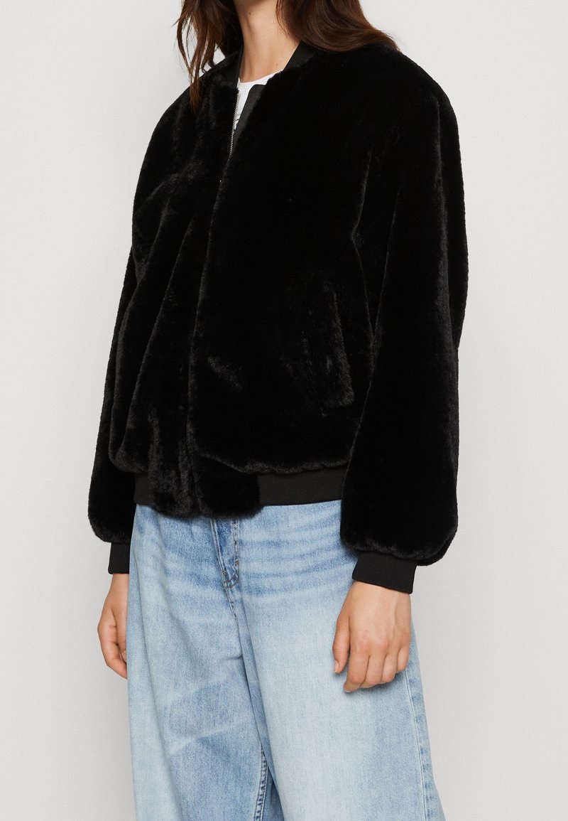 Sort faux fur bomberjakke med lynlås foran, ribbede manchetter og kant. Båret over en hvid top og parret med lyseblå vide jeans.