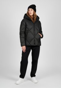 Giacca nera imbottita con motivo a diamante, cappuccio, zip frontale e tasche laterali, abbinata a pantaloni neri e sneakers bianche.