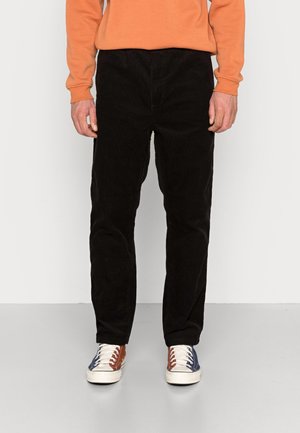 Persona con pantaloni neri, maglia arancione a maniche lunghe e scarpe da ginnastica multicolore in piedi su un pavimento grigio contro una parete bianca.