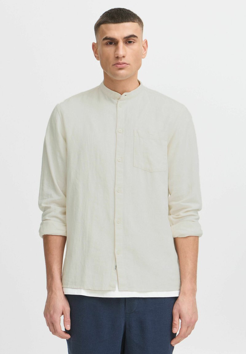Camisa de lino en un tono beige claro, con cuello alzado, cierre de botones y un bolsillo en el pecho, con mangas remangadas.
