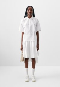 Hvit polo skjorte med korte ermer, med perforerte detaljer, lagd over en plissert hvit kjole. Modellen holder en beige håndveske.
