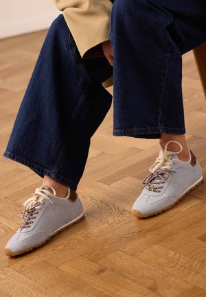 Person wearing beige coat and dark blue wide-leg jeans sitting, showing white suede sneakers with patterned laces on wooden floor.