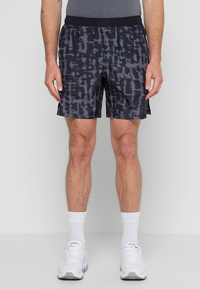 Homme portant un short de sport noir à motif, des chaussettes blanches de type crew et des baskets blanches, se tenant contre un fond gris clair uni.