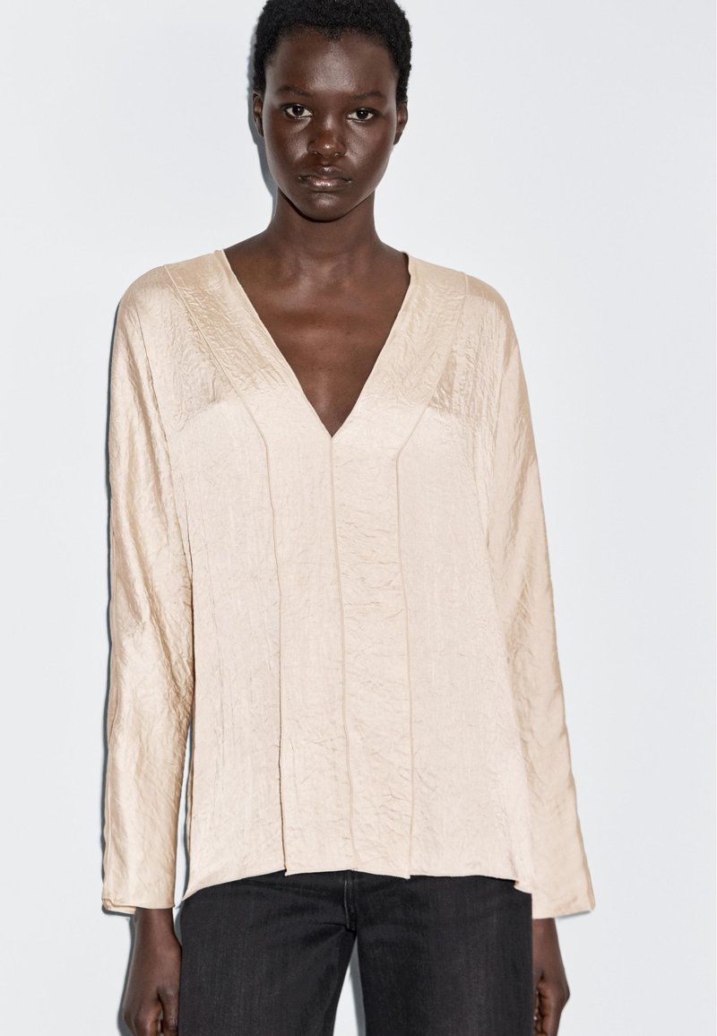Beige, lightweight long-sleeve top with a deep V-neck, textured fabric, and vertical seam detailing; paired with dark jeans.