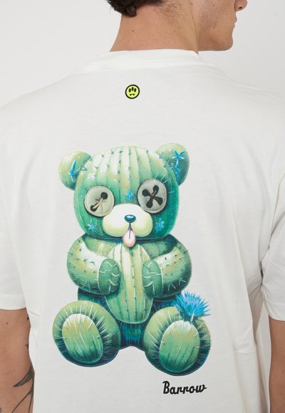 Uomo che indossa una camicia bianca con un orsacchiotto verde a forma di cactus che tiene un cactus e un fiore blu stampati sulla schiena.