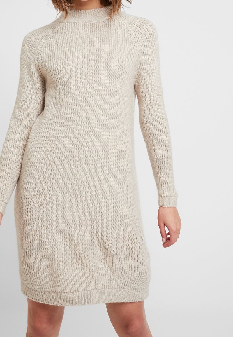 Femme portant une robe pull tricotée beige à manches longues et longueur genoux, debout devant un fond clair uni.