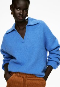 Blauer Wollpullover mit Kragen und V-Ausschnitt, der eine weiche Textur aufweist. Kombiniert mit braunen Cordhosen.