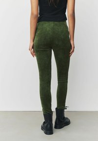 Pantalon en velours côtelé vert taille haute avec une texture lisse et une jambe fuselée, associé à des bottines noires à lacets.