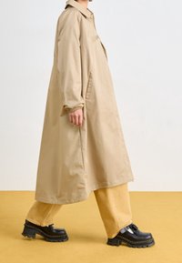 Béžový dlouhý kabát s límcem, elastickými manžetami a bočními kapsami, doplněný světle žlutými kalhotami a masivními černými botami na platformě.