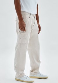 Hombre con pantalones cargo beige claro con múltiples bolsillos y zapatillas blancas con suelas de goma, de pie contra un fondo liso.