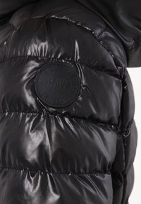 Sección de chaqueta acolchada negra con textura brillante y acolchada. Parche de logo redondo prominente con "G-STAR RAW" estampado en la manga.