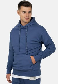 Homme portant un sweat à capuche bleu avec poche avant et une chemise blanche superposée en dessous, regardant de côté avec la main dans la poche.