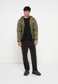 Jachetă puffer de culoare verde măslin, cu fermoar și glugă, purtata cu un tricou negru și pantaloni texturați negri. Are adidași negri.