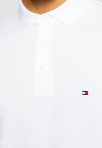 Polo shirt blanc en tissu texturé, avec un col à trois boutons et un petit logo brodé en bleu, rouge et blanc sur la poitrine.