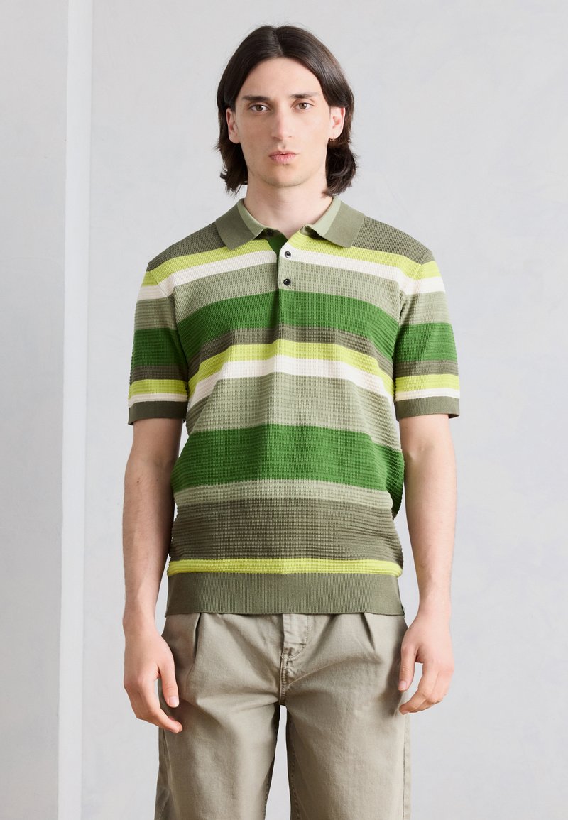 Jeune homme aux cheveux foncés mi-longs portant un polo rayé vert à manches courtes et un pantalon beige sur fond uni.