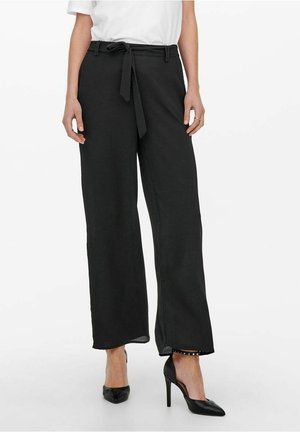 Frau trägt schwarze Hosen mit weitem Bein und einem Stoffgürtel, der in der Taille gebunden ist, kombiniert mit schwarzen High-Heel-Schuhen und einem weißen Oberteil.