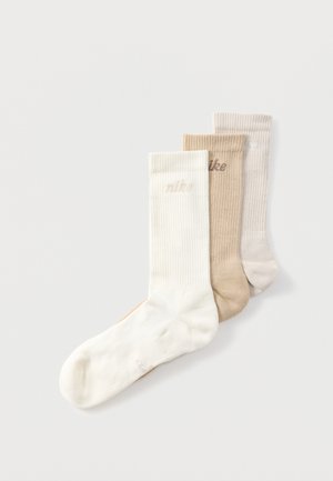 Tre foldede knæsokker i hvid, beige og lys tan med et subtilt "nike"-logo på hver manchet, arrangeret overlappende på en hvid baggrund.
