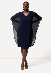 Abito di chiffon blu notte con scollatura a V e maniche a cappuccio morbide e fluide. Presenta un tessuto traslucido e un orlo arrotondato. Indossato con tacchi con cinturini.