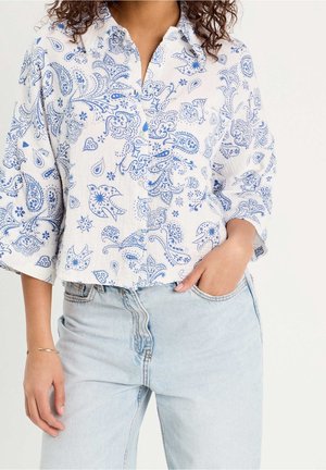 Frau trägt ein weißes Hemd mit blauem Paisley- und Vogelmuster, eine hell gewaschene Jeans, eine Hand in der Tasche, stehend vor einfarbigem Hintergrund.