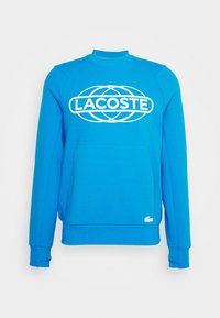Blue sweatshirt featuring a white Lacoste logo, crew neck, long sleeves, and a front kangaroo pocket. Soft fabric with ribbed cuffs.
